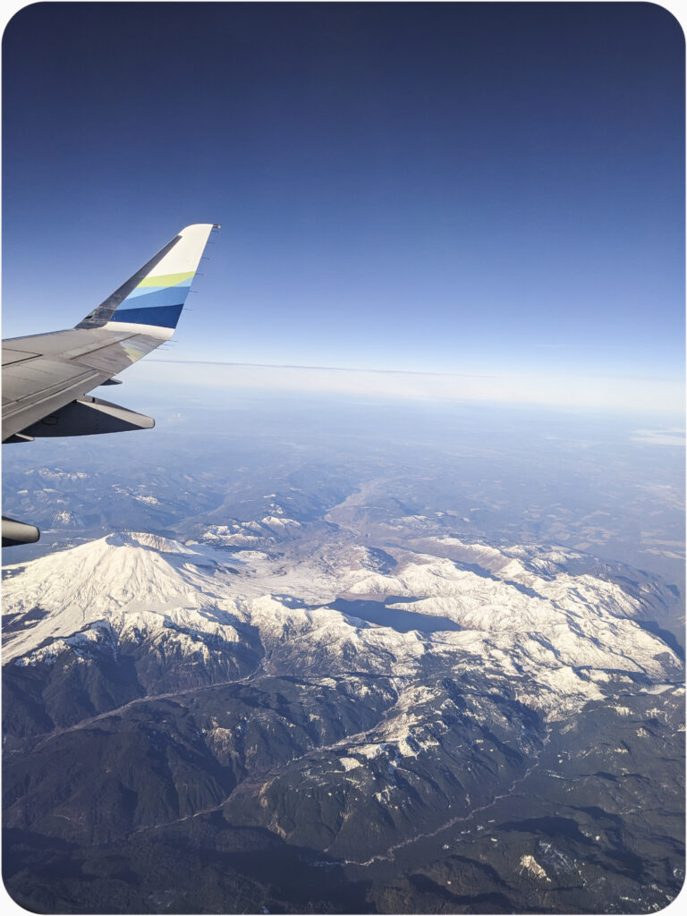 A view of Mt St Helens as captured from the right side of an airplane. The wing is visible which means the overhead compartments were full by the time they called my boarding group. Mt St Helens is covered in snow which is blueish in color.