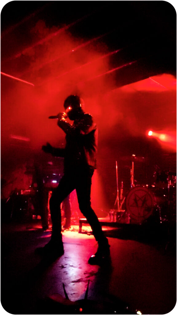 Interior of a music venue. The dark room and stage are illuminated by red light. The red light, in turn, is broken by a figure in black, singing into a microphone. There is a glowing red light emanating from the singer's face.