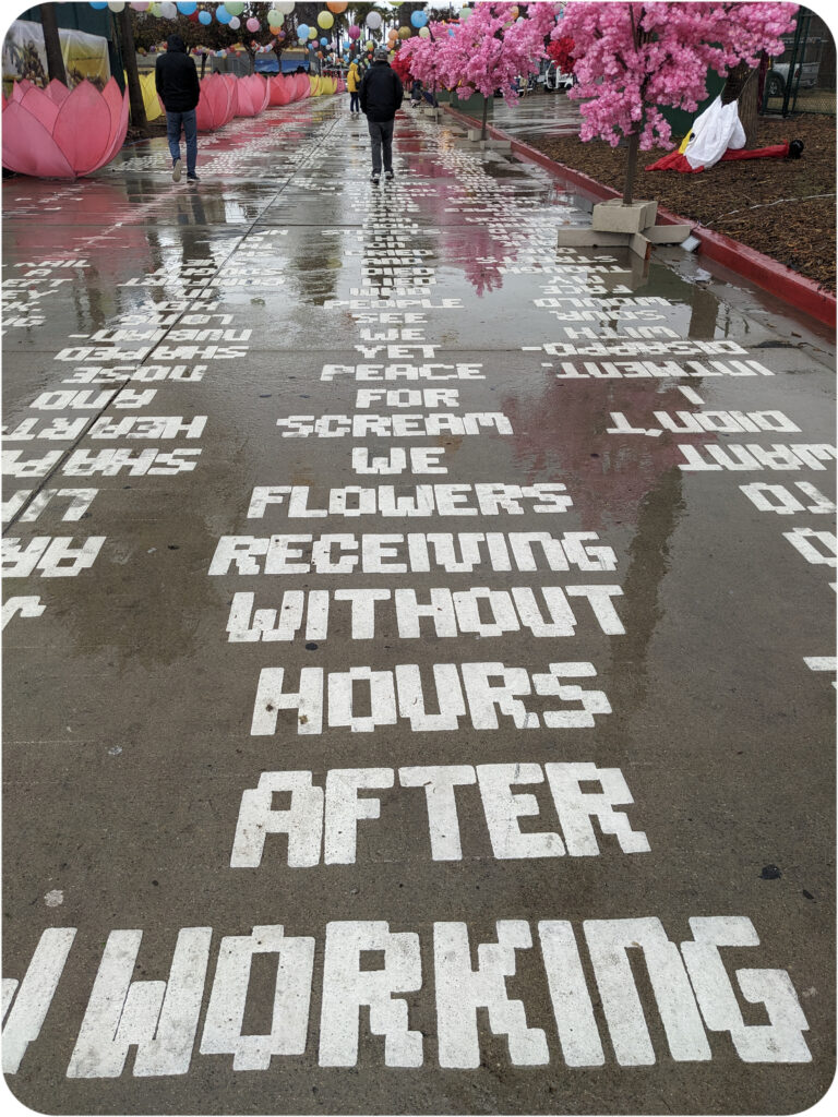 The wet painted roadway leading to the new year festival. The words read "Working after hours without receiving flowers we scream for peace yet we see people who lied died and cried on the streets how can we have peace when their stomachs" the piece continues past the gaze of the camera. 