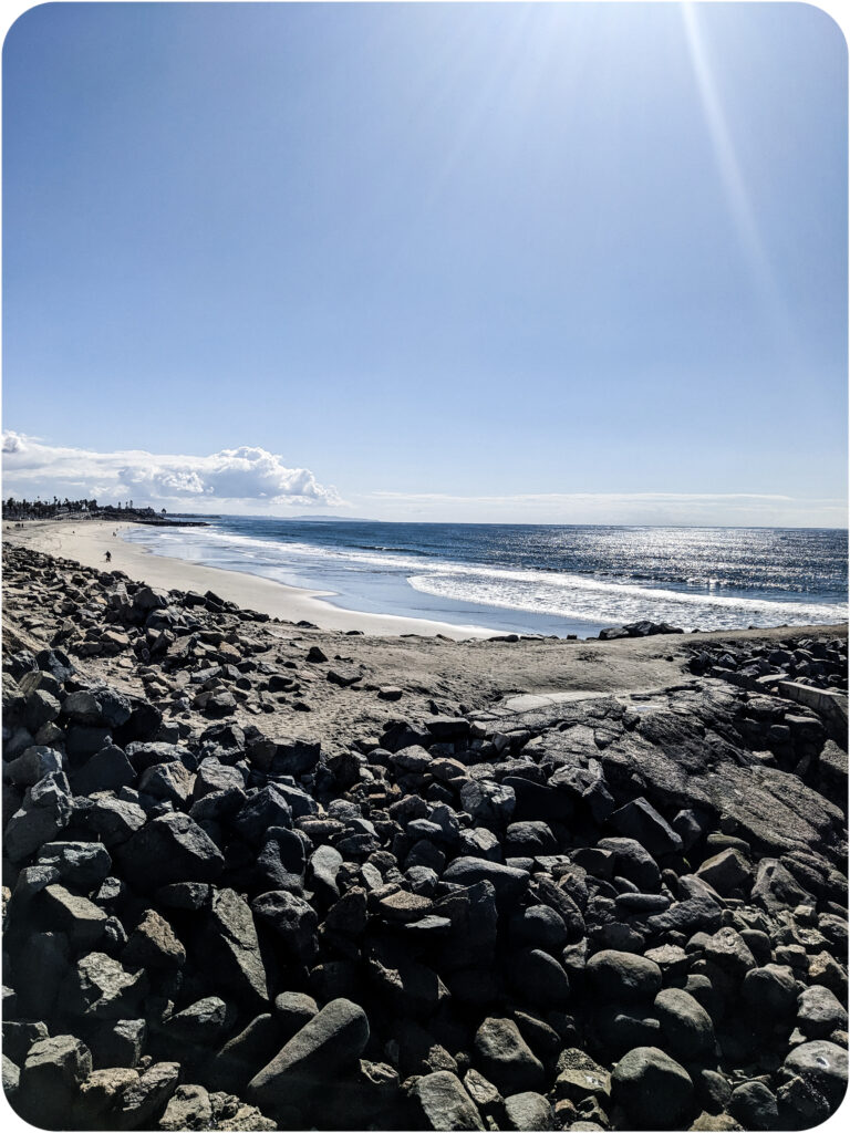 A viewpoint along Carlsbad Beach Boulevard. The foreground is textured by slate grey angular rocks. The middle ground is the sand of the beach, the light blue of the surf, the intense dark blue of the ocean and above, a bright, nearly cloudless blue sky.