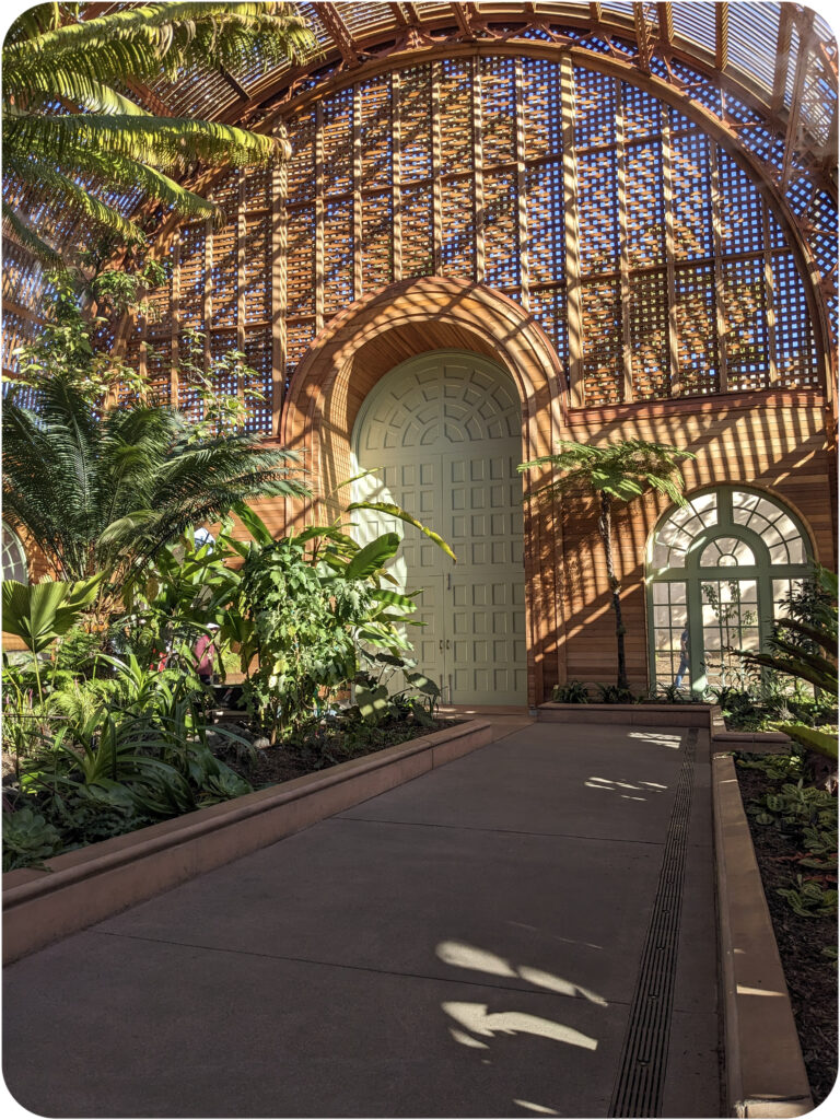 The interior of the Balboa Botanical Building. The curve of the roof is echoed by the curve of the inset heavy wood door, which has been carved and painted white. The curve is echoed again by the ground windows of the structure. Although the window is large, perhaps as many as ten feet at its apex, it appears small when the context for it is an enormous door, perhaps 15 feet at its apex.