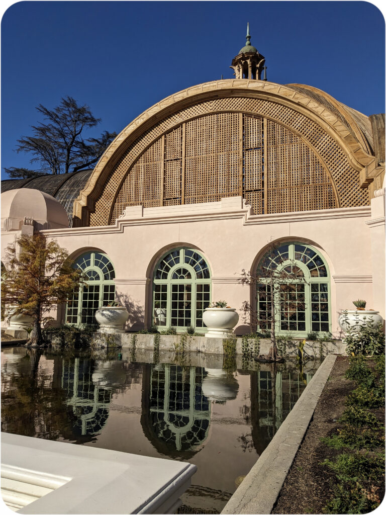 The exterior of the balboa park botanical building. In the late afternoon light, the wood domes of the roof appear lightly golden. The exterior is a soft pink stucco, with three large arched windows, green with verdigris. These windows are reflected in the reflecting pool beneath them.