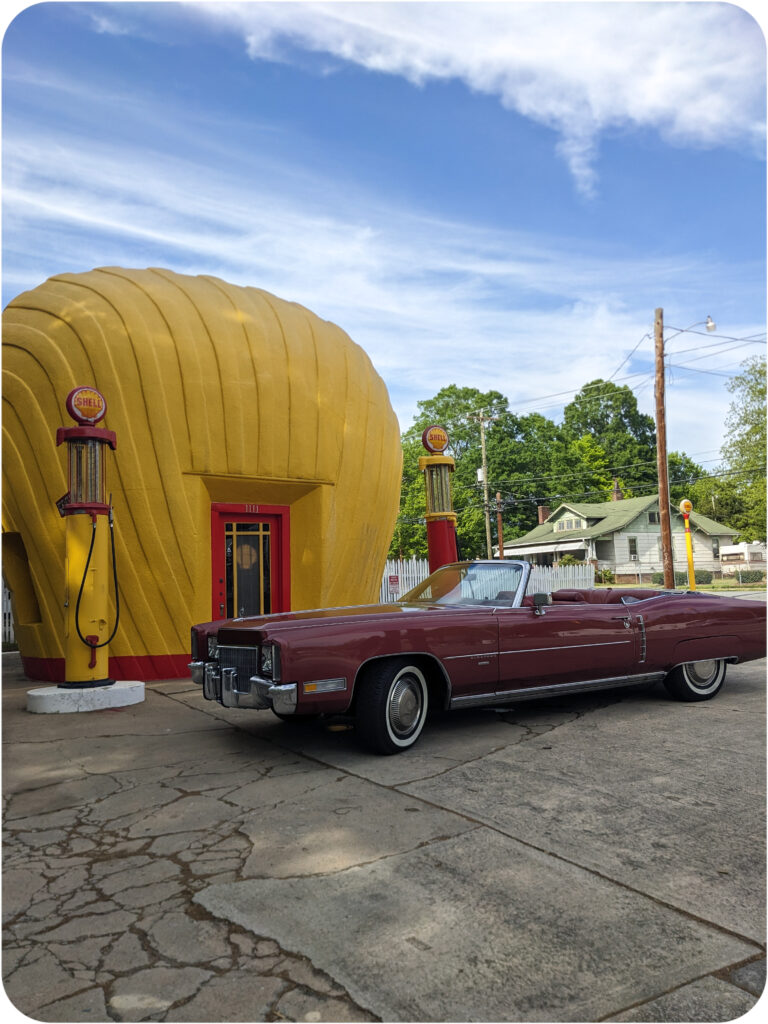 A Shell gas station that is shaped like a large yellow clamshell. A burgandy Cadillac El Dorado is parked in front.